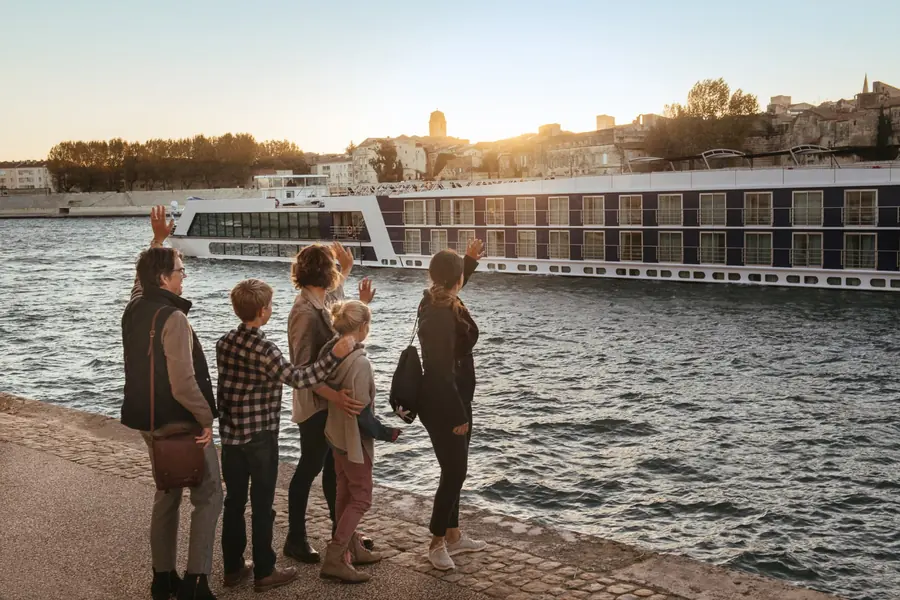 A family waving from the riverbank as a river cruise ship passes at sunset